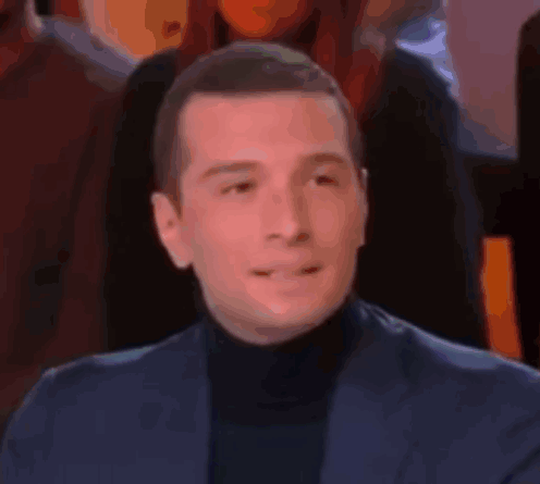 a man in a suit and turtleneck is sitting in front of a crowd of people .