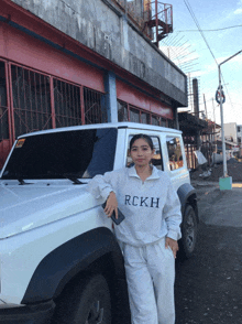 a woman wearing a sweatshirt that says rckh stands next to a white suv