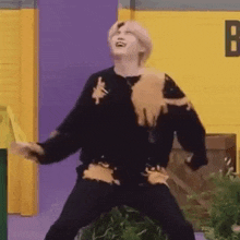 a man in a black sweater is standing in front of a yellow wall and laughing .