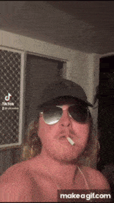 a shirtless man wearing sunglasses and a baseball cap smoking a cigarette