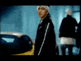 a man wearing a bandana and a black jacket is standing in front of a yellow car .