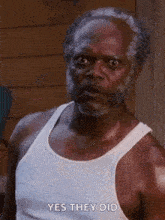 a man with a beard is wearing a white tank top and looking at the camera .