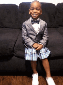 a young boy wearing a suit and bow tie is sitting on a black couch
