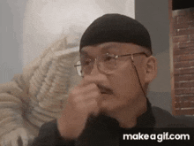 a man wearing glasses and a hat is covering his nose with his hands .