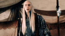 a man with long blonde hair and a crown on his head stands in front of a tent