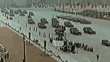 a row of cars are driving down a road with people standing on the sidewalk