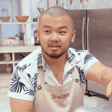 a man with a shaved head and a beard is wearing an apron that says sheldon