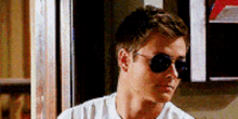 a man wearing sunglasses and a white shirt is standing in front of a sign that says ' a ' on it .
