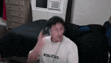 a man is sitting on a bed wearing headphones and a white shirt with the word athletics on it .