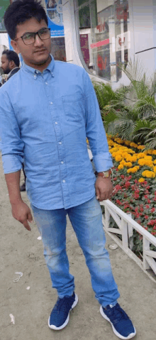 a man wearing glasses and a blue shirt is standing in front of a flower bed