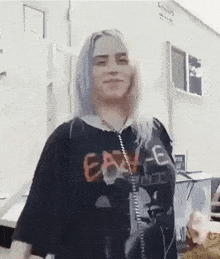 billie eilish is standing in front of a trailer wearing a black t-shirt and smiling .