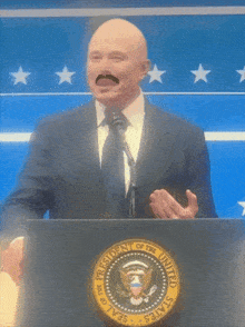 a bald man with a mustache is standing at a podium with the seal of the president of the united states