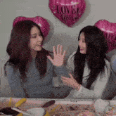 two women giving each other a high five in front of a balloon that says love you