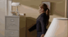 a woman in a suit is standing next to a filing cabinet and a lamp .