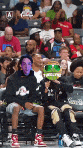 a group of people sitting in a stadium watching a basketball game and one of them has the word thedjxnz on his head