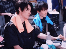 a man with a blue towel around his neck sits next to a man with an orange towel around his neck