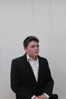 a young man in a black suit and white shirt sits with his hands folded