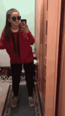 a girl wearing sunglasses and a red jacket takes a selfie in a hallway