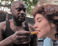 a man in a black tank top is feeding a woman a french fry