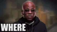 a bald man wearing sunglasses and a black hoodie is standing in front of a sign that says where .