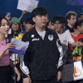 a man wearing a gen.g jacket stands in a crowd of people
