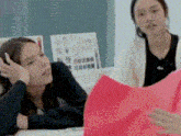 two women are sitting at a table looking at a piece of paper . one of the women is holding a red piece of paper .