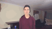 a young man is standing in a living room wearing a baseball cap and a maroon hoodie .