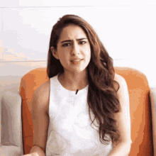 a woman with long hair is sitting on an orange couch making a funny face .