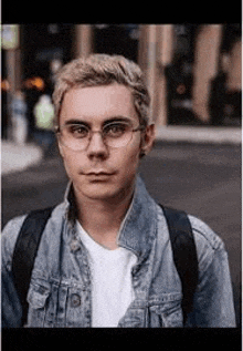 a young man wearing glasses and a denim jacket is standing in front of a building .
