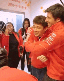 a man in a red jacket with chinese writing on the sleeve is hugging another man .