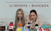 two women are standing in front of microphones in front of a wall that says lumphini bangkok