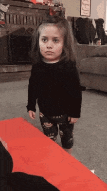 a little girl is standing on a red mat in a living room with a serious look on her face .