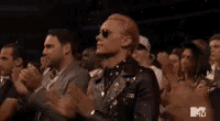 a man with pink hair and sunglasses is standing in a crowd of people clapping .