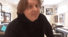 a man in a black hoodie is standing in a living room with a piano and guitars on the wall .