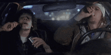 two men are sitting in a car at night . one of the men is smoking a cigarette .