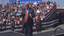 donald trump stands on a stage in front of a crowd with signs that say america great again