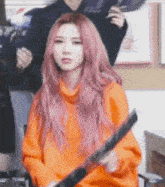 a woman with pink hair is wearing an orange sweater and holding a sword .