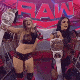 two female wrestlers holding their championship belts in front of a raw sign