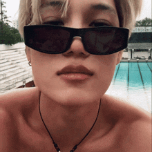 a woman wearing sunglasses and a necklace stands in front of a swimming pool