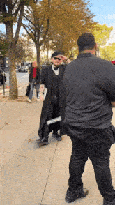 a man wearing sunglasses stands on a sidewalk next to a man wearing a black shirt that says " supreme "