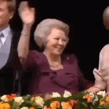 a woman in a purple dress is sitting in front of a table with flowers and waving her hand .