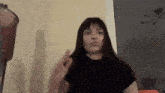 a woman in a black shirt is making a funny face and waving her hand in front of a window .