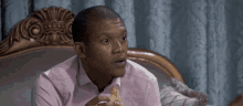 a man in a pink shirt is sitting on a couch eating a slice of pizza .