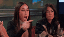a woman is pointing at another woman while sitting at a table with wine glasses .