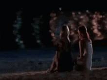 two women are sitting on the beach at night looking at each other .