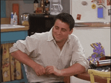 a man in a white shirt is sitting at a table in a kitchen with his hands folded .