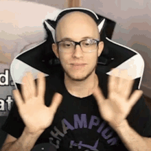 a bald man wearing glasses and a champion shirt is sitting in a chair with his hands in the air .