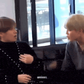 two men are sitting next to each other in front of a window and talking to each other .