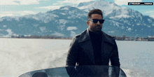 a man wearing sunglasses and a leather jacket stands in front of a lake with mountains in the background
