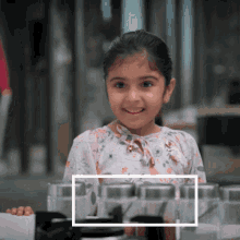 a little girl in a floral shirt stands in front of a row of containers